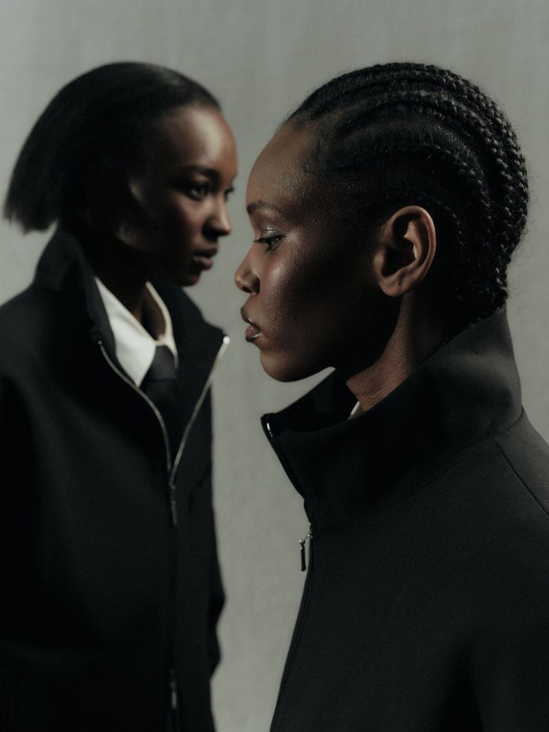 Two Black models in black high-collar zip jackets against a plain grey studio backdrop.