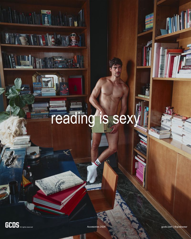 Model Alessio Pozzi standing barefoot in white socks covering himself with an open book in a library for the GCDS campaign.