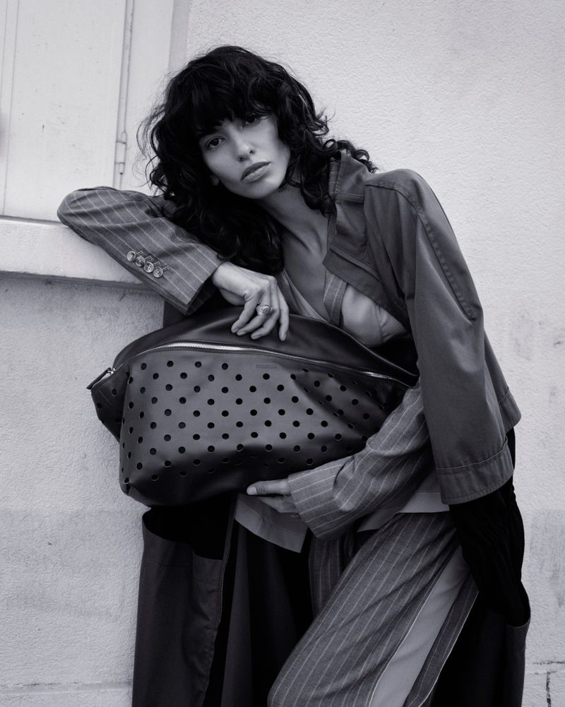 Model with dark wavy hair wearing striped blazer and trousers holding a perforated leather bag against a pale wall.