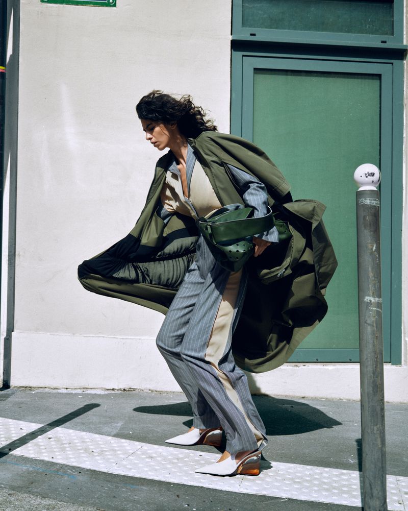 Gal Levi wearing a billowing olive green coat and grey pinstripe trousers on a city sidewalk for 10 Magazine Deutsch.