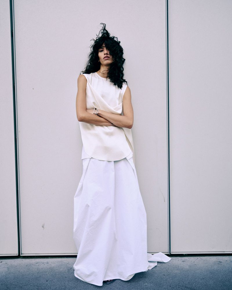 Model Gal Levi standing with arms crossed wearing a sleeveless cream top and long flowing white skirt against a paneled wall.
