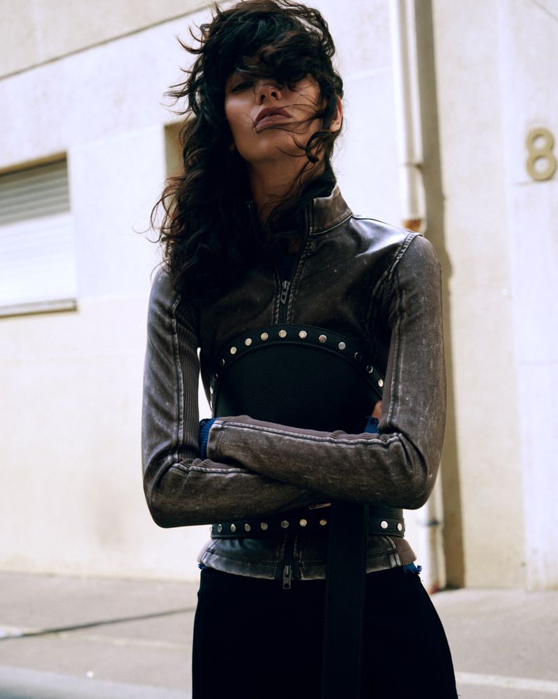 Model with windblown hair wearing a distressed brown leather jacket and studded corset belt outdoors.