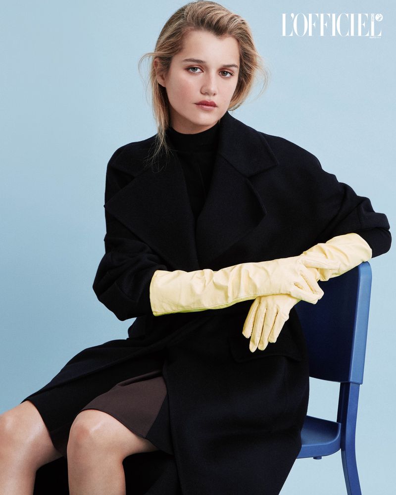 Fernanda Finsterbusch seated wearing an oversized black coat and long pale yellow gloves against a light blue backdrop.
