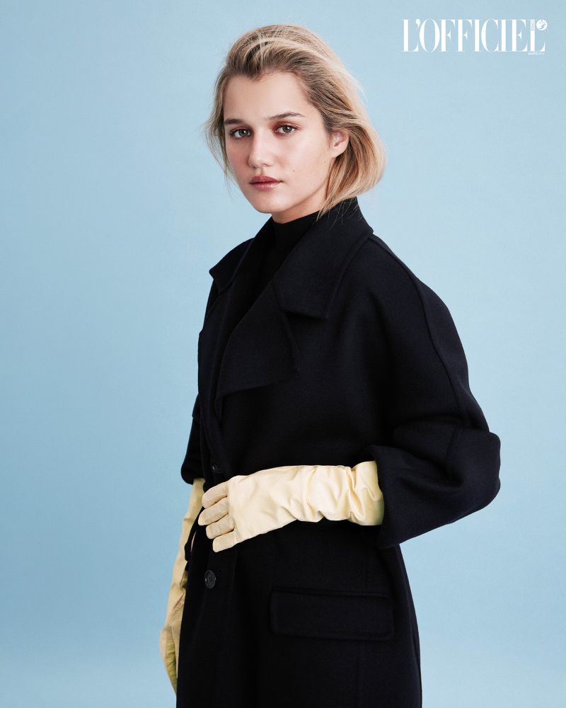 Fernanda Finsterbusch in an oversized black coat and pale yellow leather opera gloves against a light blue seamless studio backdrop.