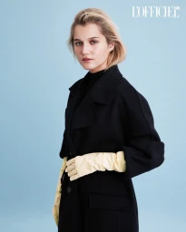 Fernanda Finsterbusch in an oversized black coat and pale yellow leather opera gloves against a light blue seamless studio backdrop.