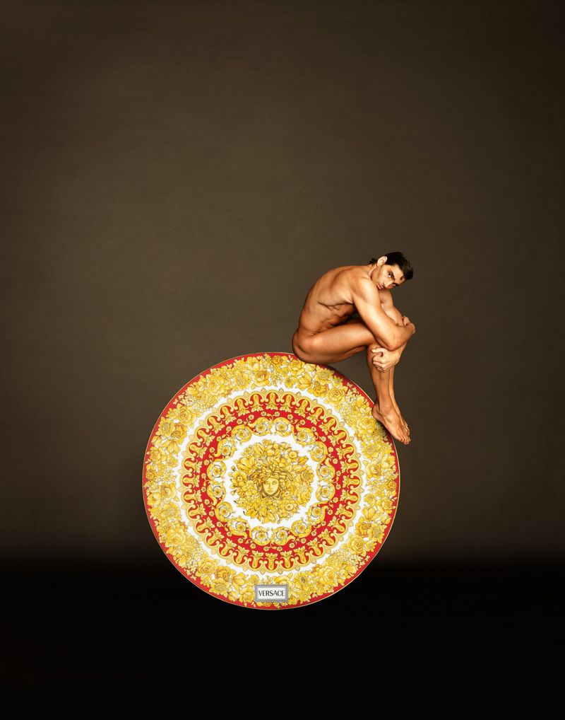 Nude male model crouching atop a large ornate gold and red circular Versace medallion platter against a dark background.