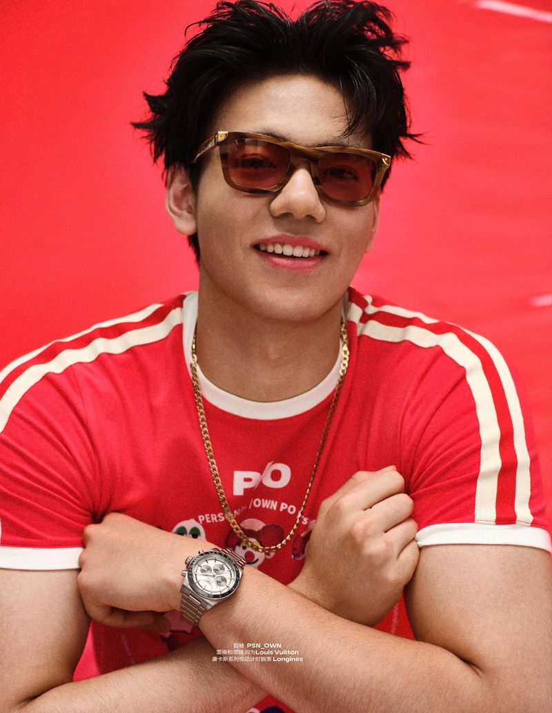 Young East Asian man in tinted sunglasses and a red graphic T-shirt smiling with arms crossed against a red backdrop.