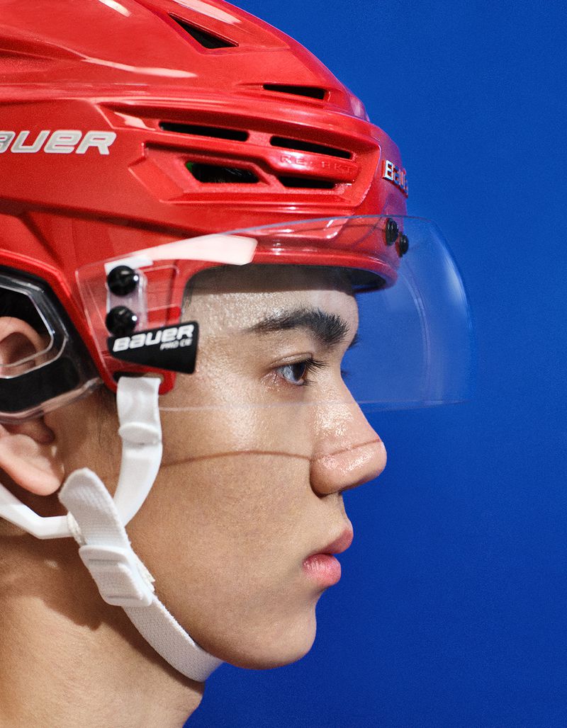 Close-up profile of a young East Asian man wearing a red hockey helmet with a clear visor against a blue background.