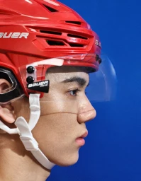 Close-up profile of a young East Asian man wearing a red hockey helmet with a clear visor against a blue background.