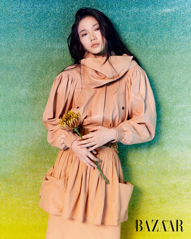 Model Ching Yeung wearing a peach puffed-sleeve ensemble and holding a flower against a green textured backdrop.