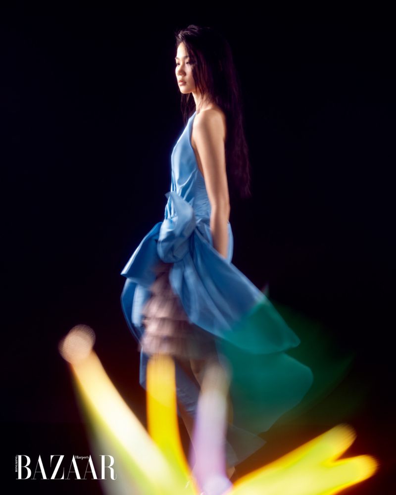 Model Ching Yeung poses in a flowing blue halter gown against a black backdrop with blurred yellow lights in the foreground.