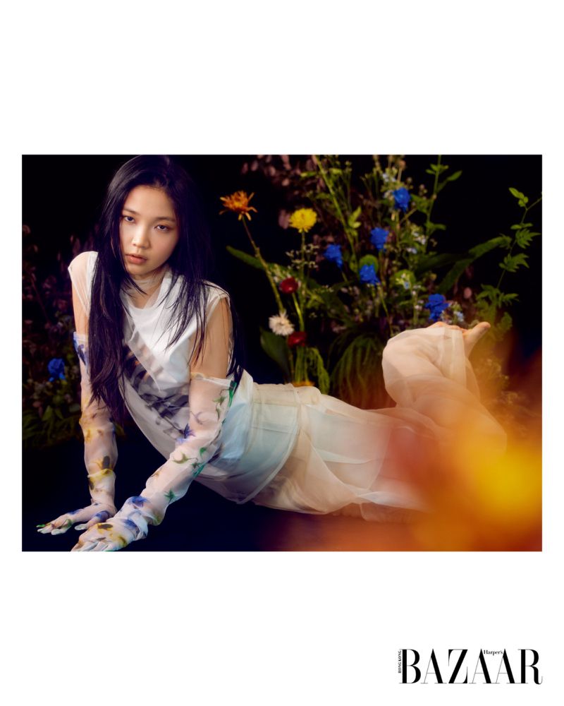 Model Ching Yeung reclining in a sheer white outfit with printed floral sleeves against a dark floral studio backdrop.