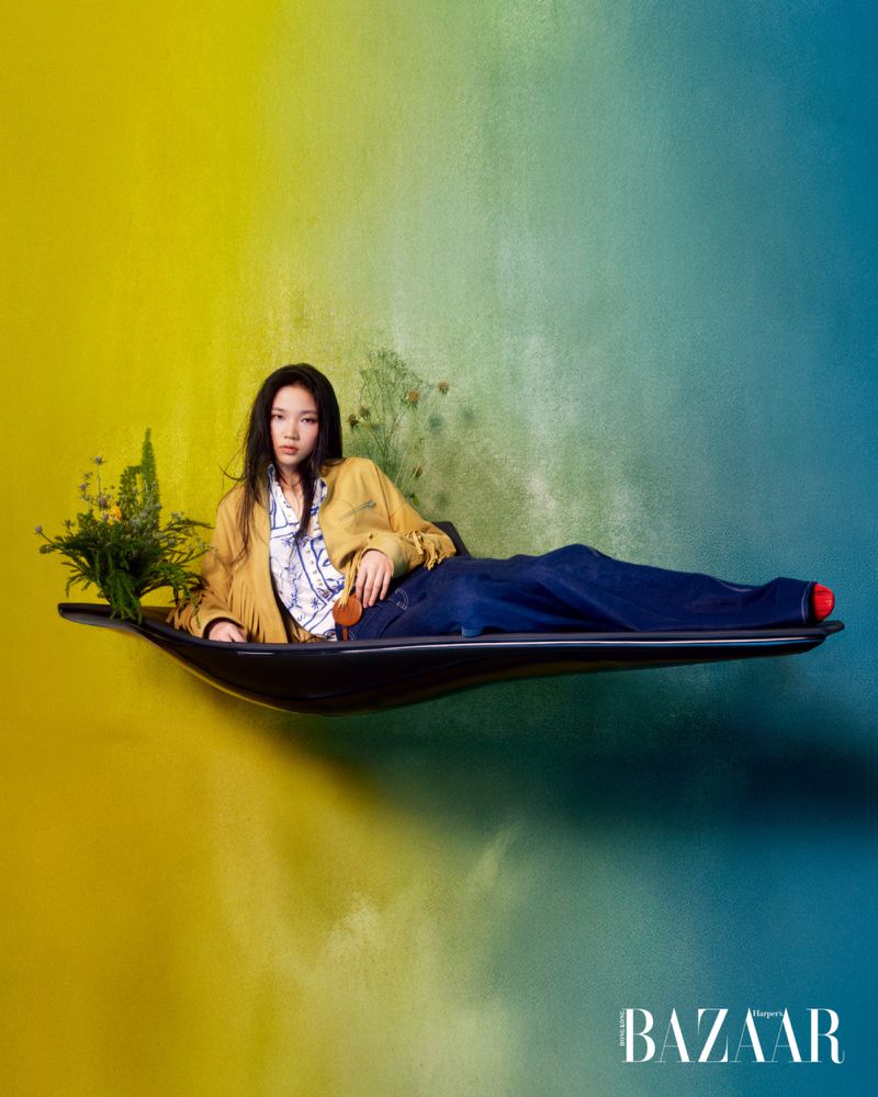 Model Ching Yeung reclining on a floating black curved bench against a yellow and blue gradient backdrop for Harper's Bazaar Hong Kong.