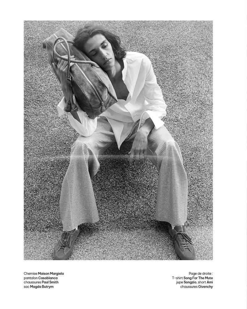 Model resting head against a large leather shoulder bag while seated outdoors on textured concrete in a white shirt and wide-leg trousers.