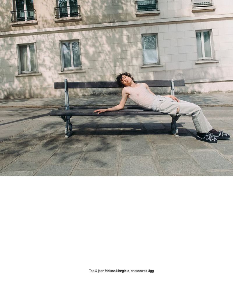 Model reclining on a wooden public bench in front of a stone building on a paved street sidewalk.