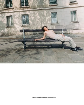 Model reclining on a wooden public bench in front of a stone building on a paved street sidewalk.