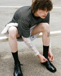 Model Laurent Batilliot seated outdoors wearing a striped top, lace sleeve, black socks, and black dress shoes while tying laces.