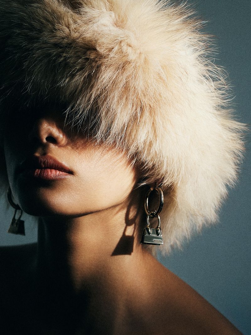 Model wearing a large fluffy beige fur hat covering her eyes and silver purse drop earrings against a dark studio backdrop.