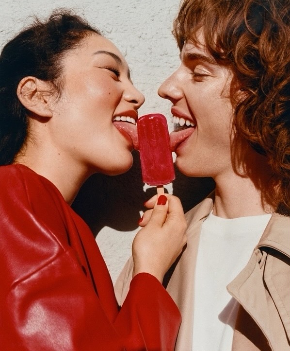 Two people, one Asian woman and one Caucasian man, sharing a red popsicle and sticking their tongues out against a neutral wall.