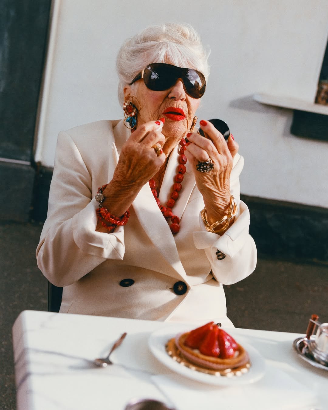 An elderly woman applies lipstick at an outdoor table, wearing large sunglasses, jewelry, and a white blazer. A tart sits on the table.