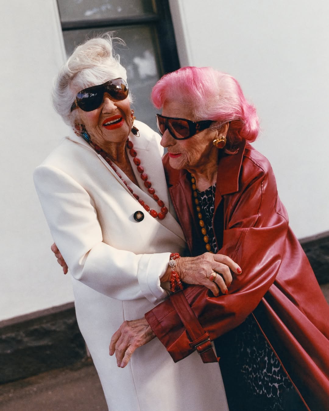 Two elderly women embrace outdoors, wearing sunglasses and stylish outfits with red beaded jewelry, standing against a light wall.