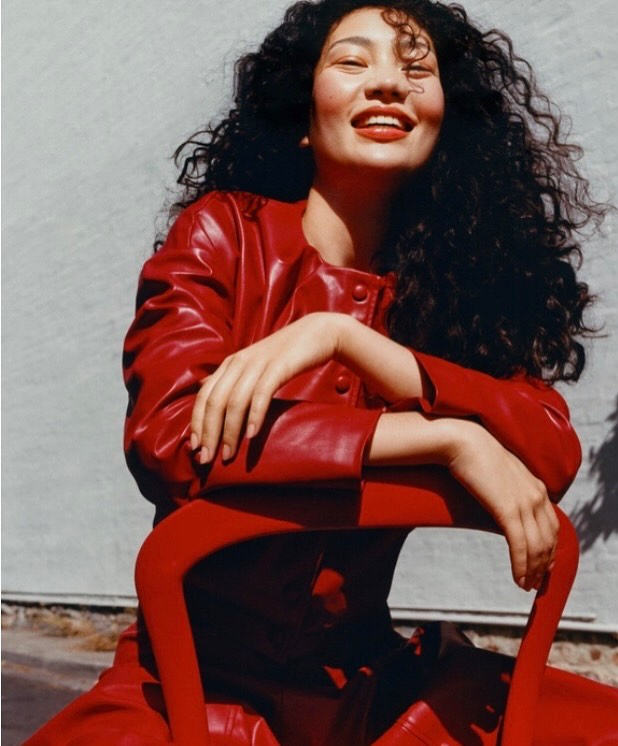 Smiling woman with dark curly hair wearing a red jacket, leaning over a red chair outdoors.