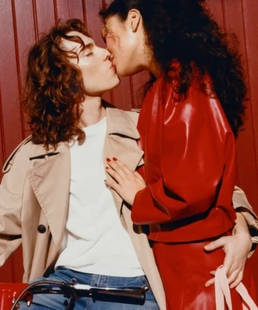 Fashion shot of a light-skinned man in a trench coat and a dark-skinned woman in a red dress kissing indoors against a paneled wall.