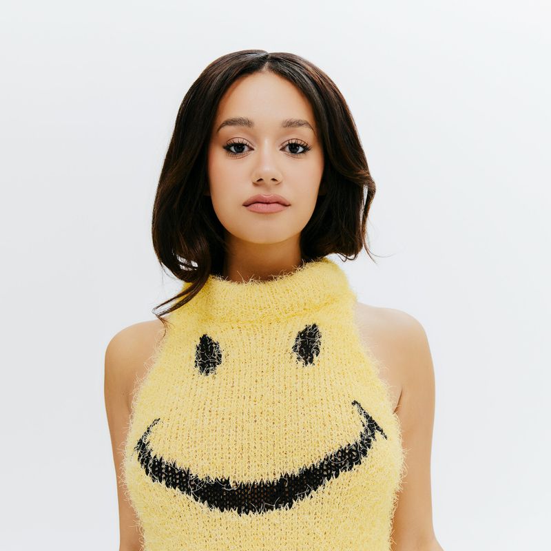 Léna Mahfouf posing in a fuzzy yellow knit halter top with a black smiley face graphic against a plain white studio backdrop.
