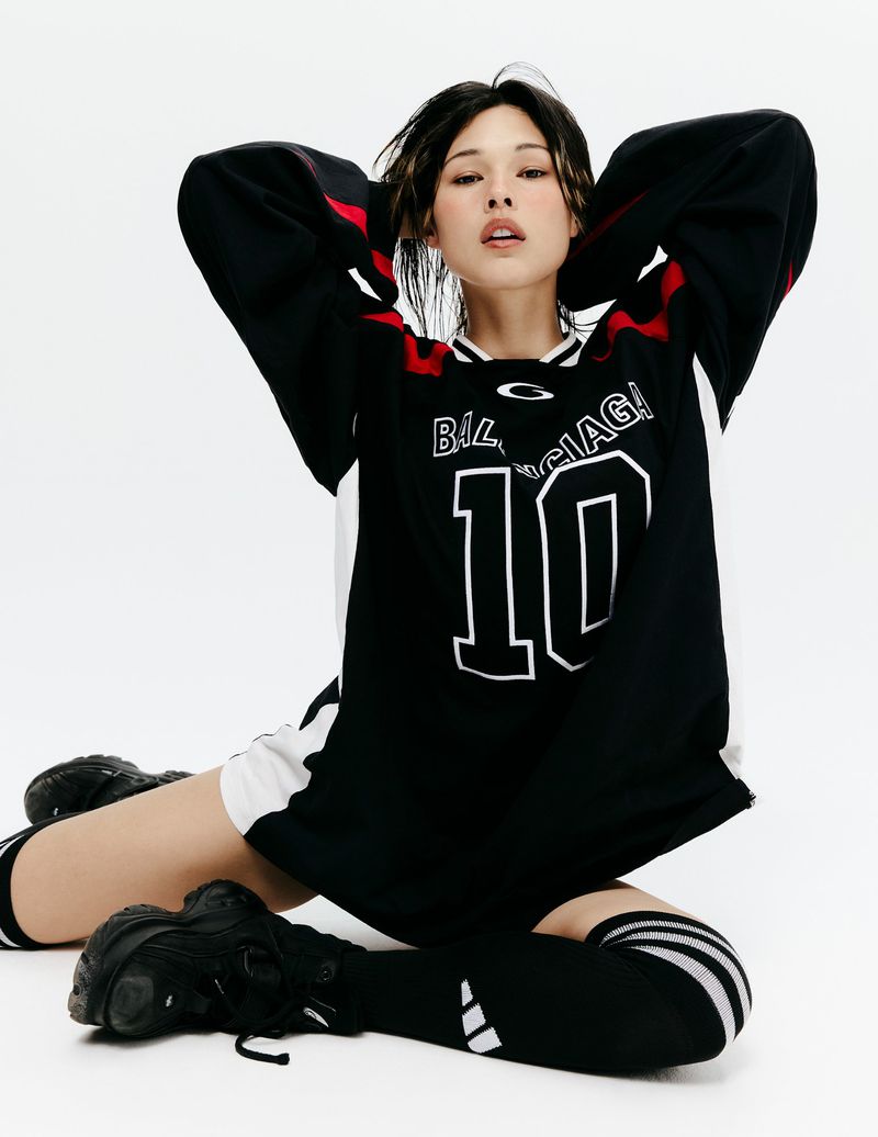 Model seated in an oversized black Balenciaga jersey, knee-high socks, and chunky black sneakers against a white background.