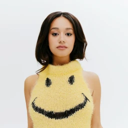Léna Mahfouf posing in a fuzzy yellow knit halter top with a black smiley face graphic against a plain white studio backdrop.
