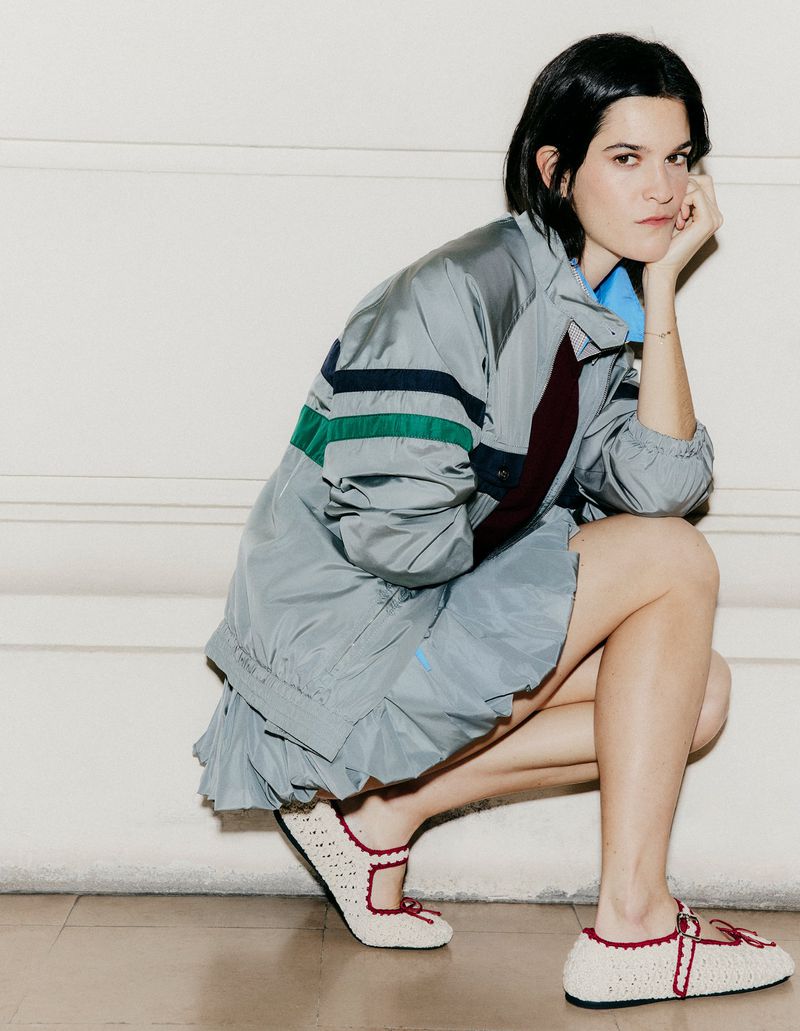 Suzanne Lindon crouching in a grey windbreaker jacket, matching pleated skirt, and cream Mary Jane flats against a neutral wall.