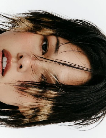 Close-up portrait of a woman with black and blonde highlighted choppy hair against a white background.
