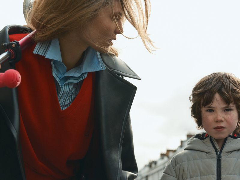 Model Ida Heiner in a black leather coat and red knit sweater looking down beside a young boy in a grey puffer jacket outdoors.