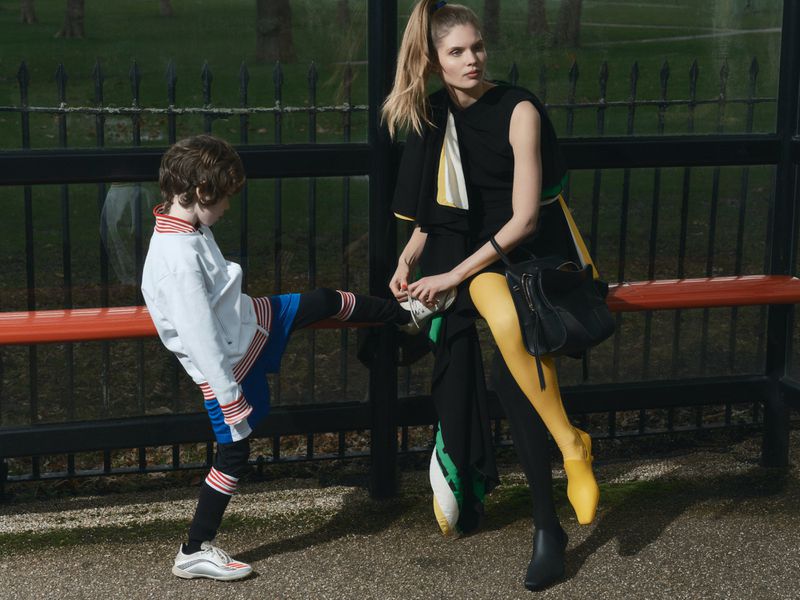 Model Ida Heiner in a black dress and yellow tights sitting at a bus stop bench beside a child stretching his leg.