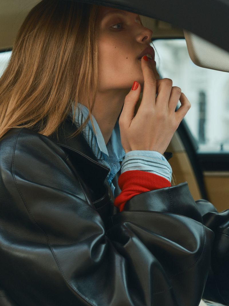 Model in black leather jacket and blue shirt sitting inside car applying red lipstick