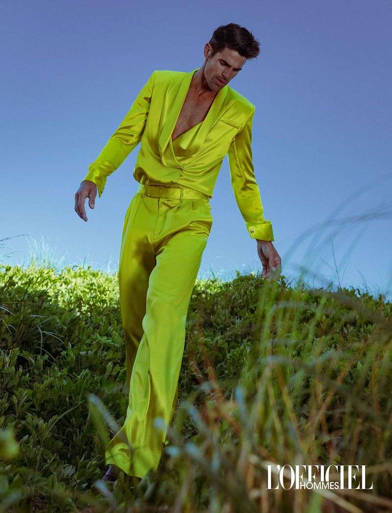 Model Chad White walking down a grassy hill wearing a bright chartreuse satin suit against a blue sky.