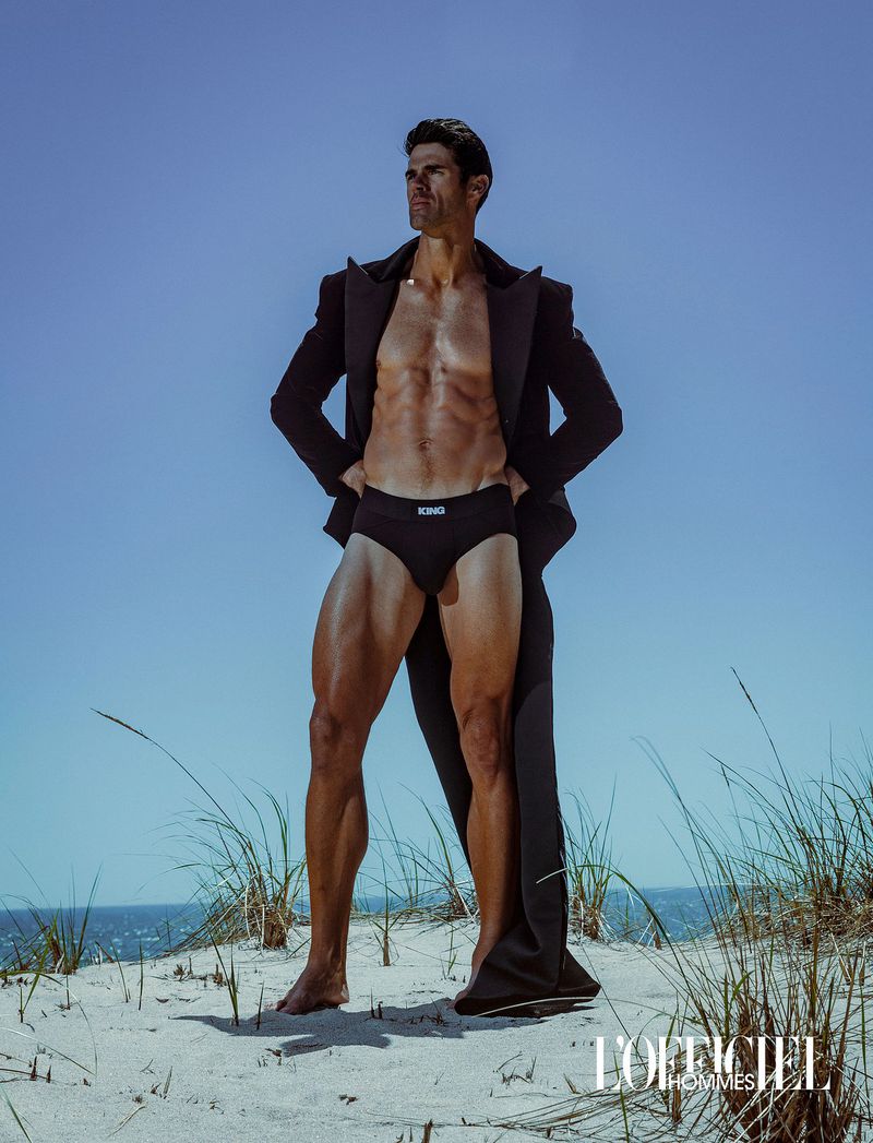 Chad White posing on a sand dune wearing an open black overcoat and black briefs for L'Officiel Hommes Ukraine.