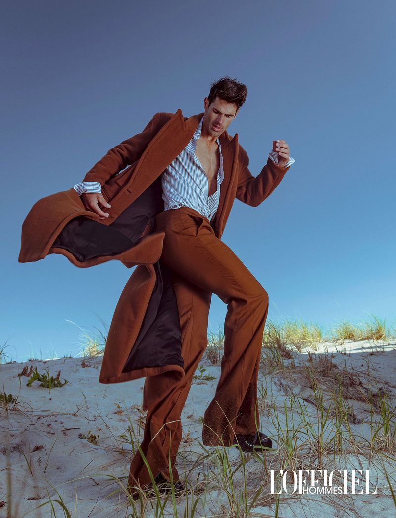 Model Chad White walking in sand dunes wearing an open brown overcoat, striped shirt, and brown trousers.