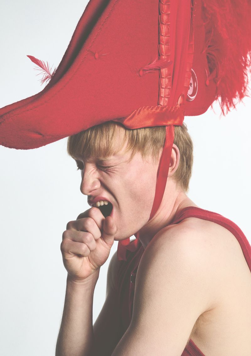 Model wearing an oversized red feathered hat and yawning against a plain white studio background.
