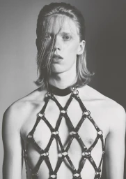 Monochrome portrait of a shirtless model wearing an intricate black leather body harness and collar against a grey studio backdrop.