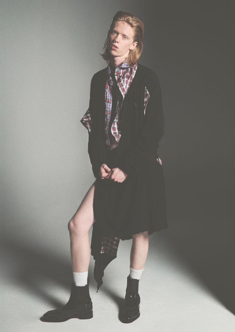 Fashion model in an oversized black cardigan, plaid shirt, white socks, and black boots posing on a grey studio backdrop.
