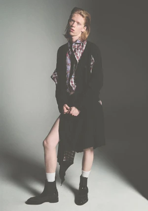 Fashion model in an oversized black cardigan, plaid shirt, white socks, and black boots posing on a grey studio backdrop.