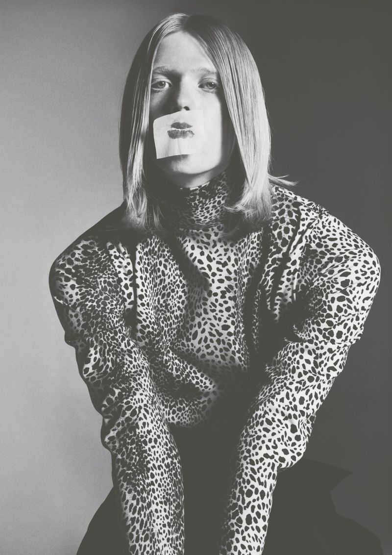 Monochrome portrait of a model wearing a leopard print turtleneck with a lipstick kiss print sticker over their mouth.