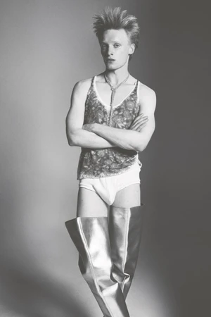 Male model with arms crossed wearing a patterned tank top, white briefs, and metallic thigh-high boots against a grey studio backdrop.