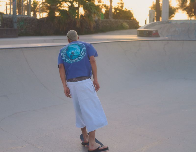 Man wearing a blue t-shirt and white shorts skateboarding in a concrete skatepark bowl at sunset.