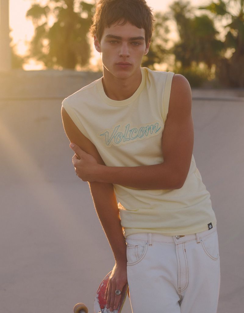 Model Jon Payeras in a light yellow Volcom sleeveless top and white jeans holding a skateboard outdoors at golden hour.