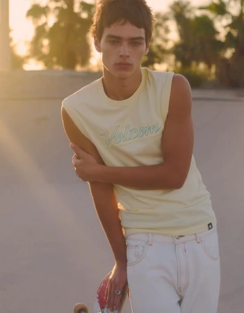 Model Jon Payeras in a light yellow Volcom sleeveless top and white jeans holding a skateboard outdoors at golden hour.