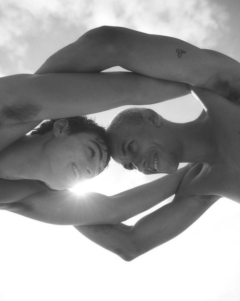 Black and white low-angle shot of two shirtless men touching foreheads with arms interlinked against a bright sky.