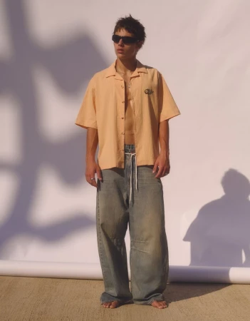 Model wearing dark sunglasses, an open peach short-sleeve shirt, and baggy denim jeans against a seamless white studio backdrop