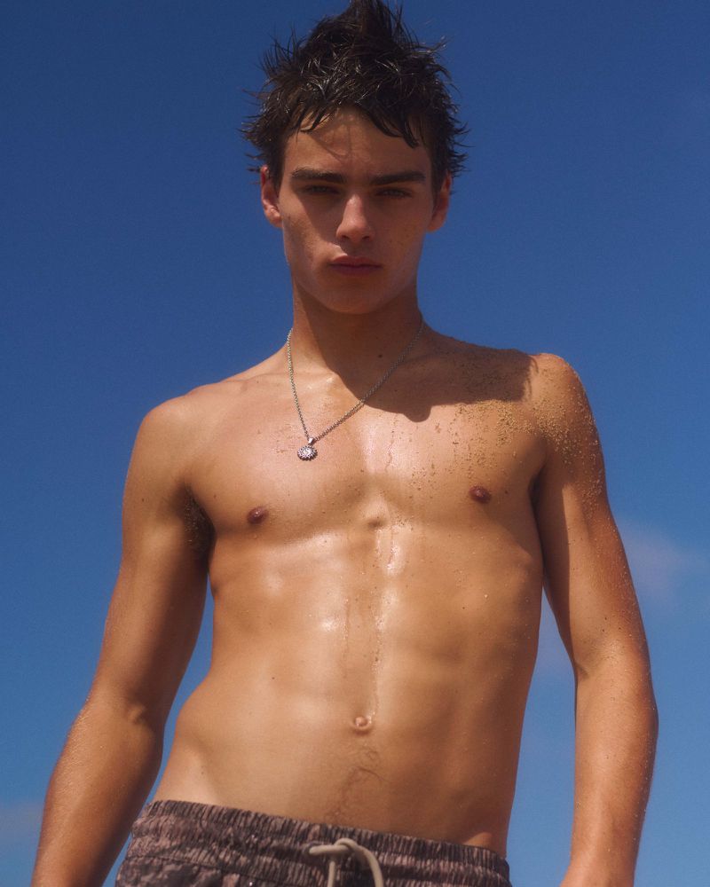 Shirtless man with wet hair and a silver necklace wearing printed swim shorts against a bright blue sky.
